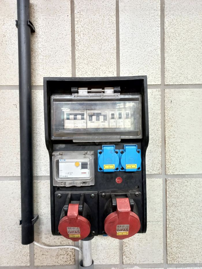 Een verdeelkast aan een betegelde muur met automaten, een teller, blauwe stopcontacten en twee rode 400 V-contactdozen.