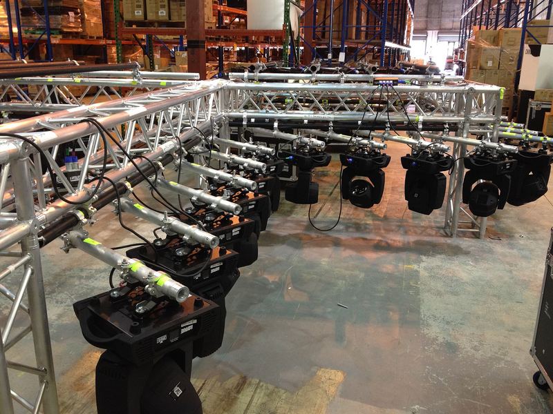 Aluminium truss op de vloer van een loods, met een rij moving heads eraan, klaar om opgetrokken te worden.