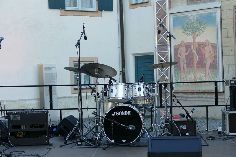 Een laag buitenpodium met leuning, gitaren, versterkers en een drumstel.