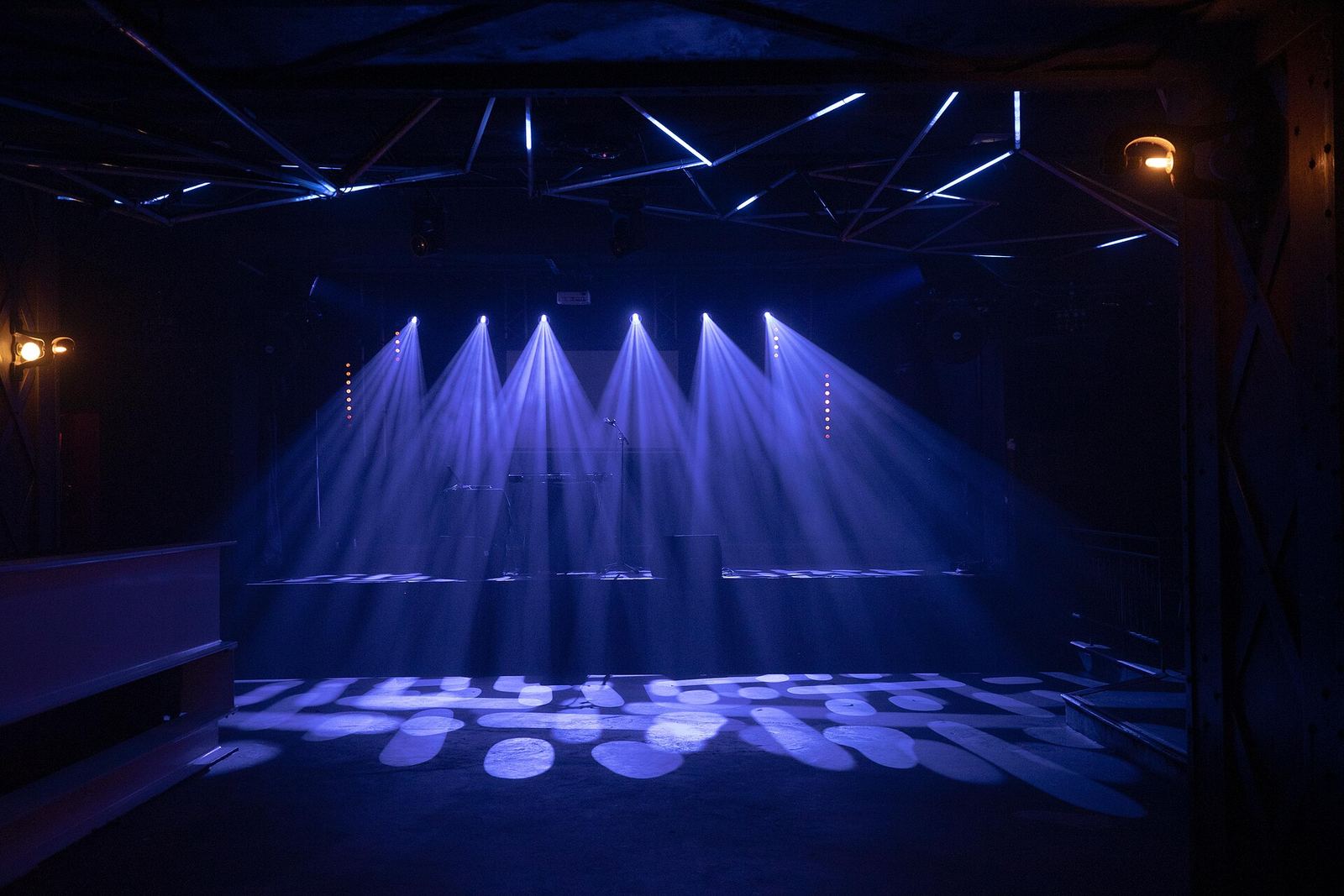 Een leeg podium in een zwarte zaal, met blauwe lichtbundels van spots aan de truss.