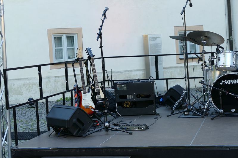 Een laag buitenpodium op een binnenplein met een drumstel, versterkers en microfoons, klaar voor een optreden.