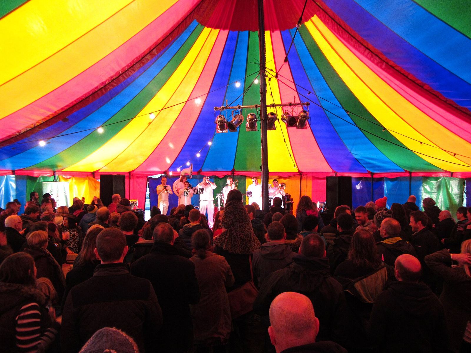 Een volle feesttent met regenboogkleurig doek, publiek en een verlicht podium achteraan.